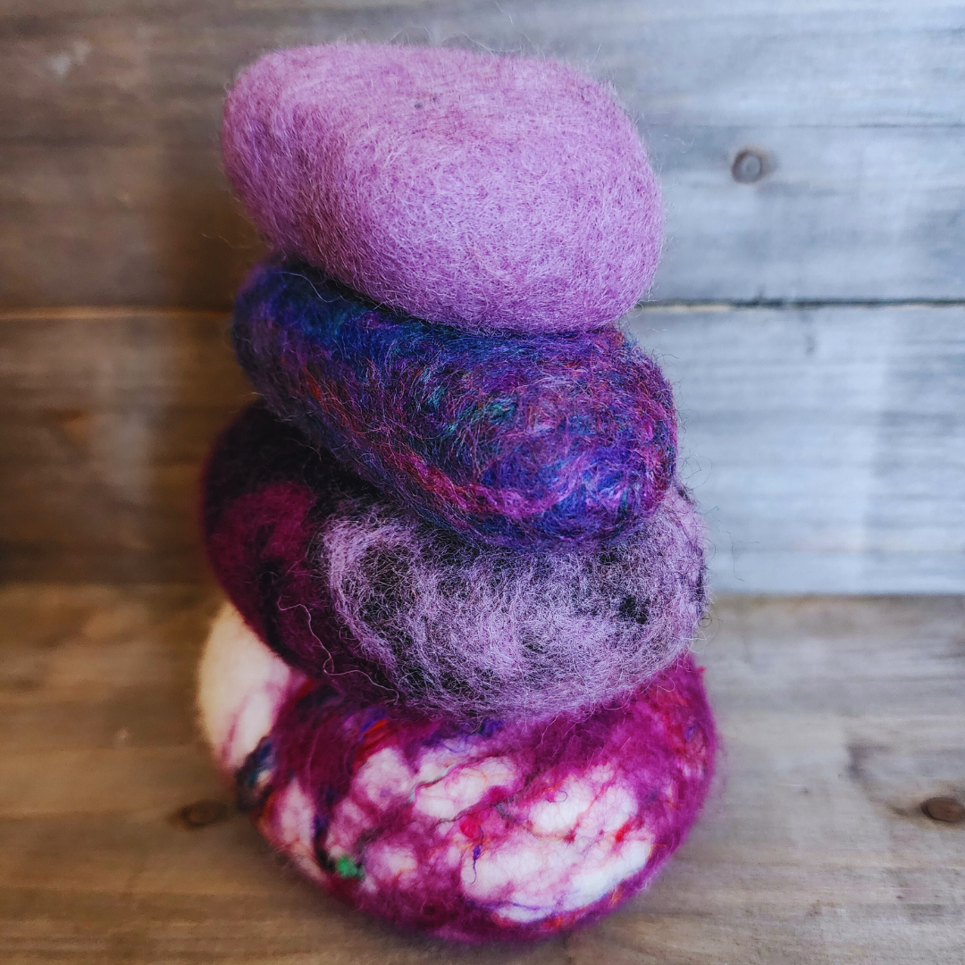 Four handmade wet-felted Zen Stones stacked in an irregular column, from bottom to top: white with pink and purple swirls, a dark purple, a blue and purple blend, and a light lavender, on a rustic wooden background.