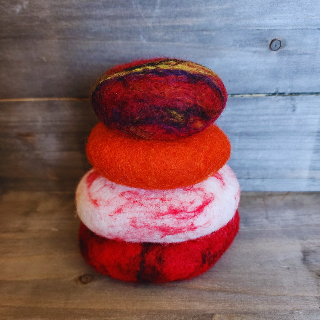 Four handmade wet-felted Zen Stones stacked in a neat vertical tower, from bottom to top: solid red, white with red swirls, solid orange, and a multi-toned top stone with red, yellow, and purple, on a wooden surface.