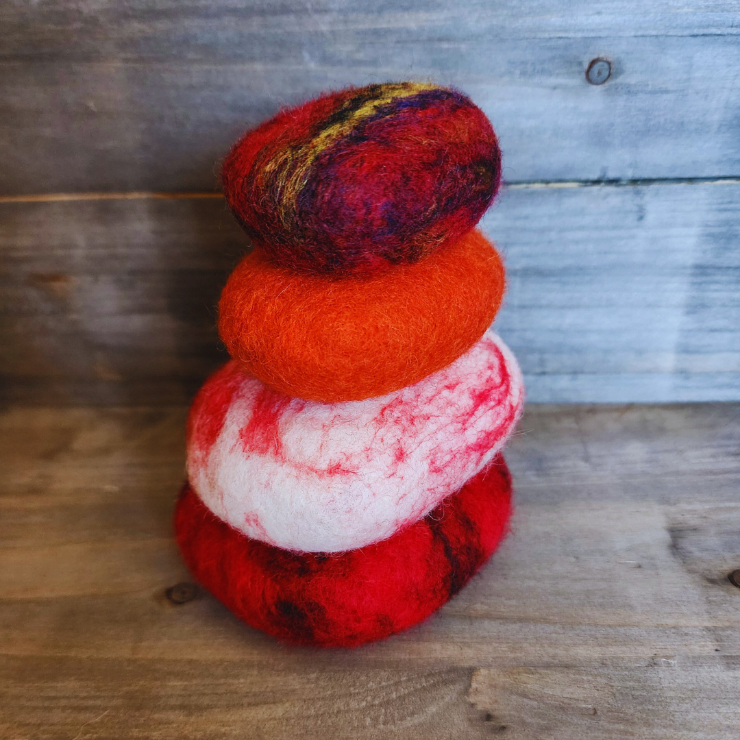 Four handmade wet-felted Zen Stones stacked vertically, featuring vibrant shades of red, orange, white with red marbling, and a multi-colored top stone with yellow and purple stripes, on a rustic wooden background.