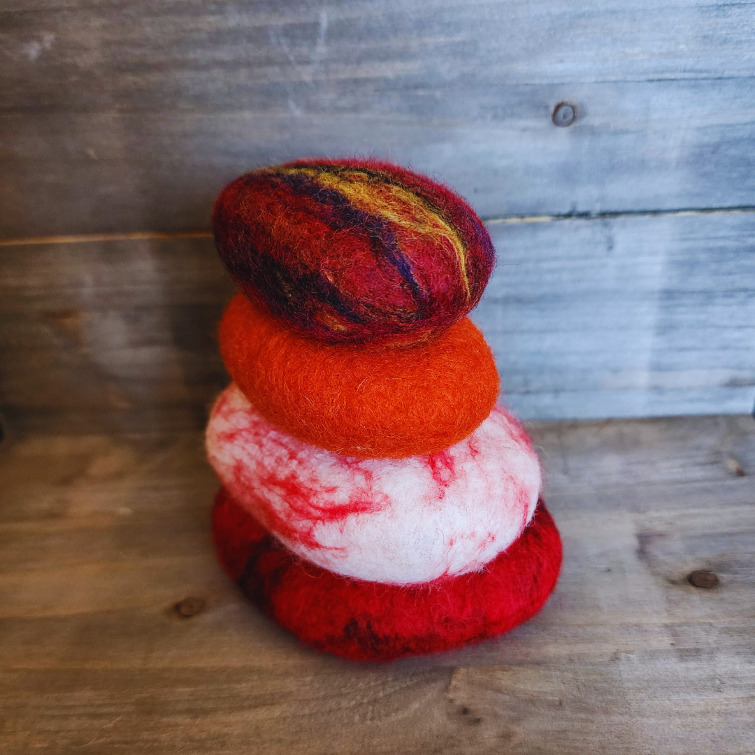 A close-up view of four handmade wet-felted Zen Stones stacked vertically, showing a bottom red stone, a white stone with red marbling, a solid orange stone, and a multi-colored top stone, against a wooden background.